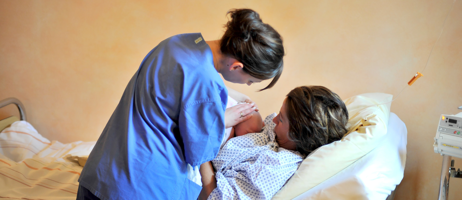 Krankenpflegerin zeigt einer frisch gebackenen Mutter, wie sie ihr Kind anlegt, damit sie es stillen kann.