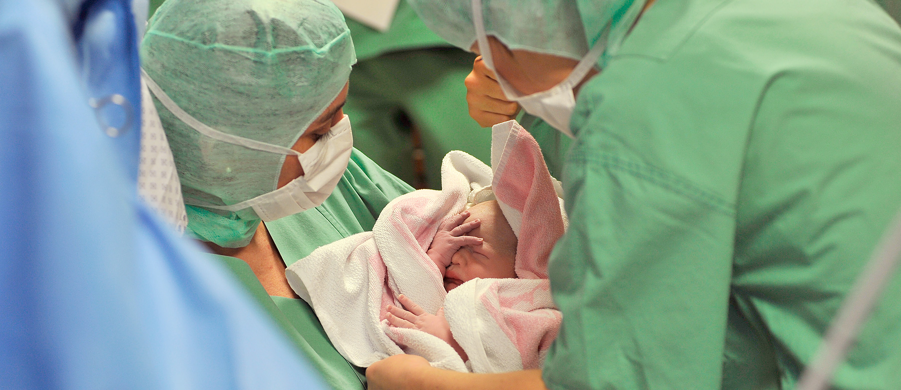 Eine Kinderkrankenpflegerin im Operationssaal hält ein Neugeborenes eingewickelt in ein Handtuch im Arm.