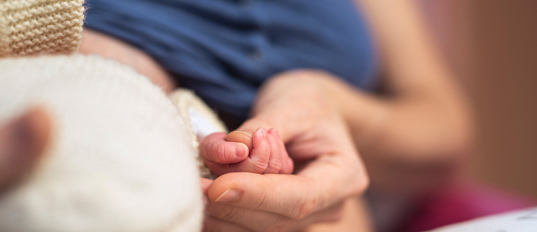 Mutter hält ihr Neugeborenes in den Armen und umgreift mit ihren Fingern die Hand ihres Kindes.