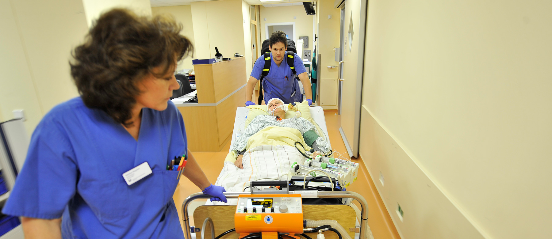 Mitarbeiterin sowie ein Notarzt schieben eine Patientin im Patientenbett über den Flur der Notaufnahme.
