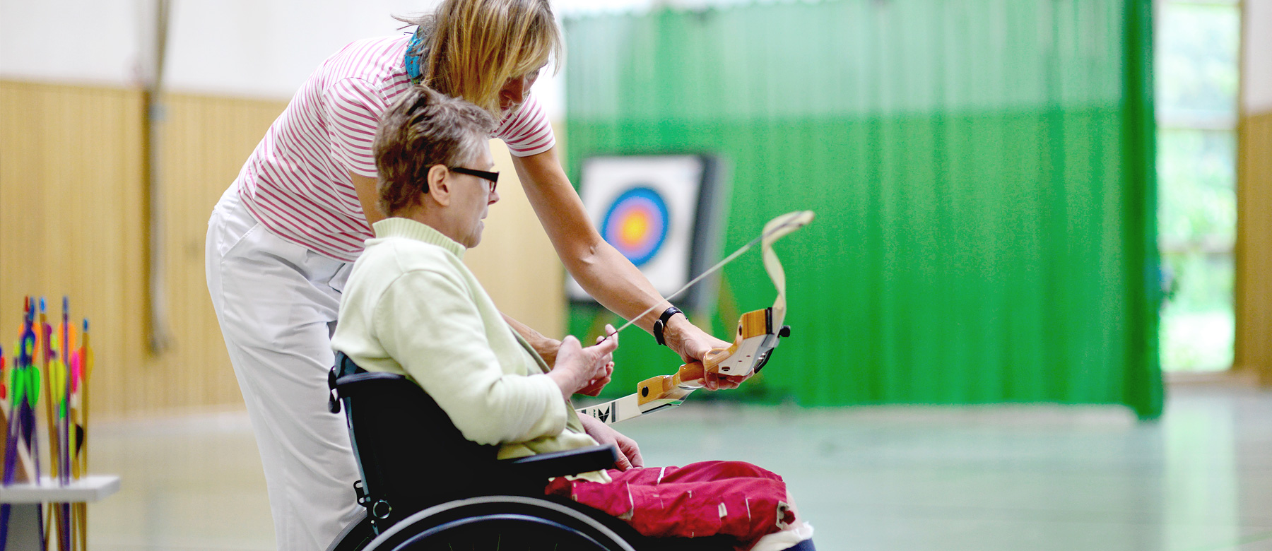 Patient im Rollstuhl beim Bogenschießen mit einer Physiotherapeutin.