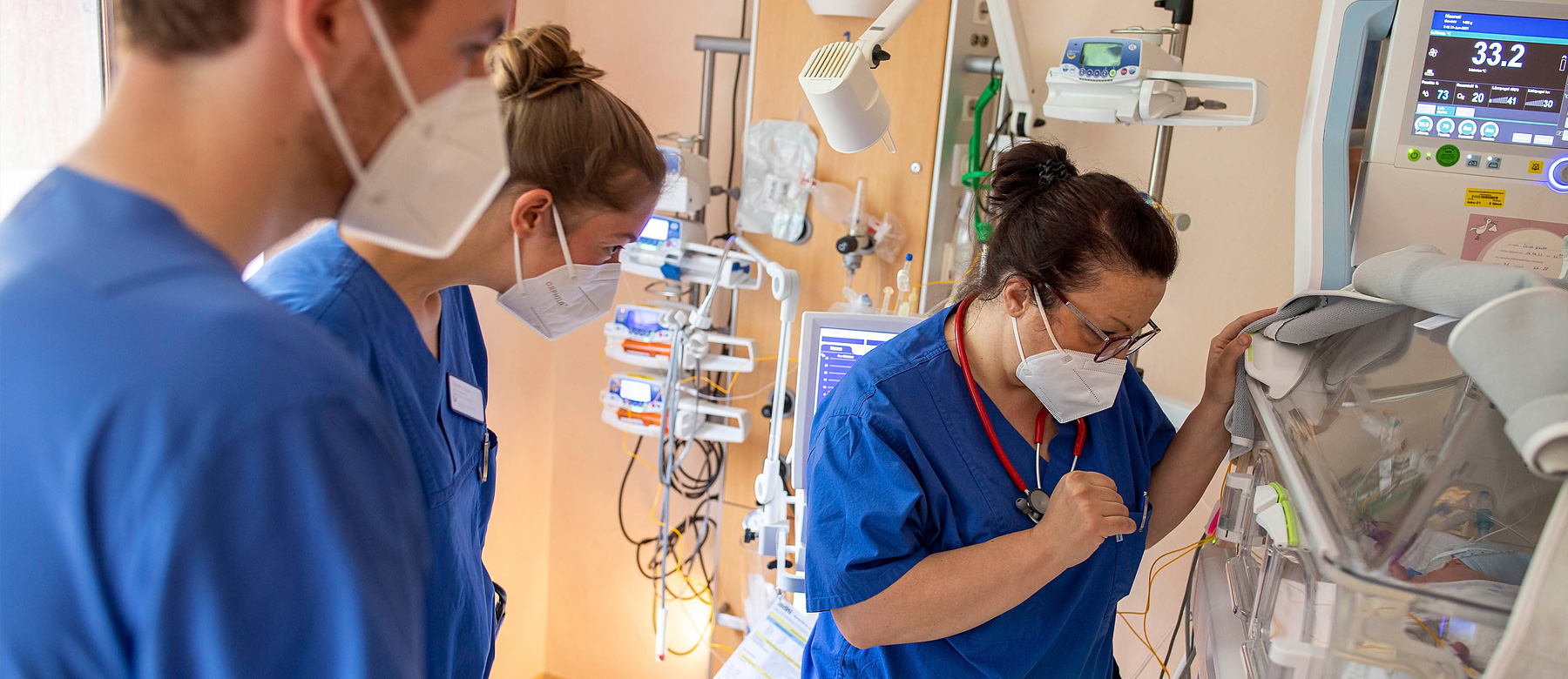 Kinderintensivpflegerin schaut nach einem Frühgeborenen im Inkubator und wird von zwei Krankenpflegeschülern begleitet.